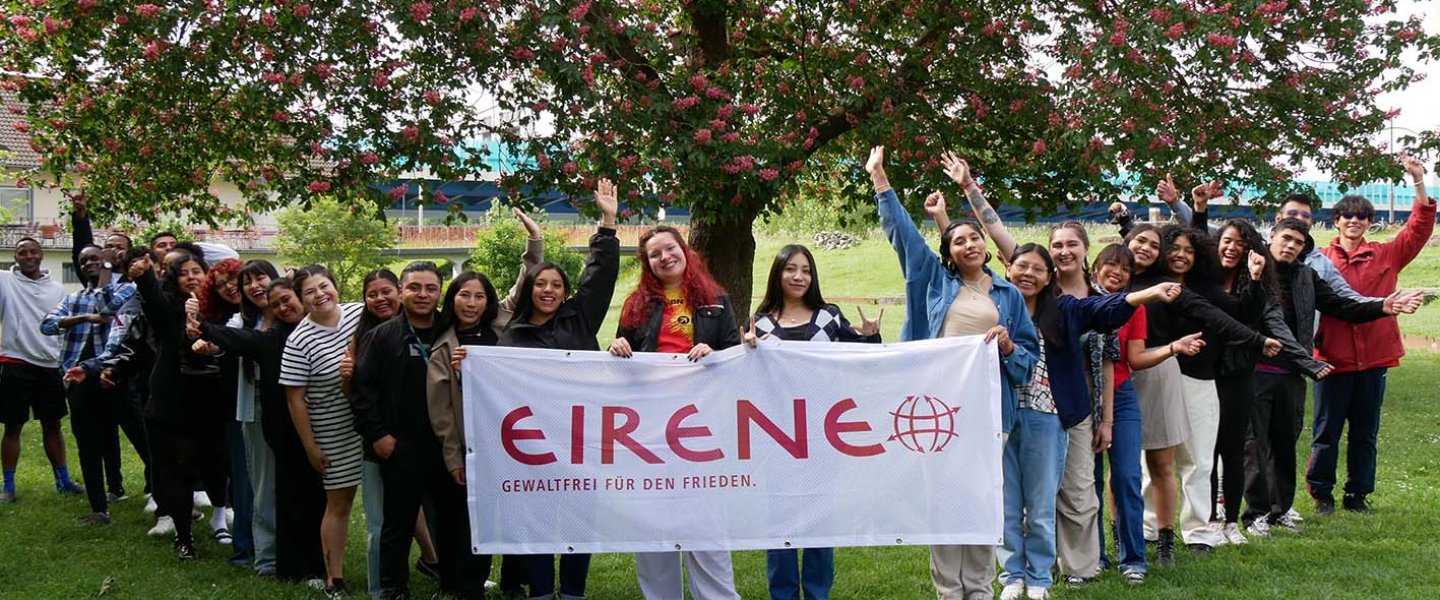 Eine Gruppe internationaler Freiwilliger steht in V-Form. Vorne tragen vier von ihnen einen EIRENE-Banner. Im Hintergrund ist ein Baum zu sehen.